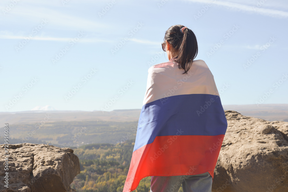 A girl tourist with a Russian flag on her shoulders stands on the top of a mountain and enjoys the beautiful views of the Caucasus Mountains.