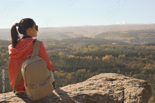 Obraz na płótnie A girl tourist with a backpack sits on the top of a mountain and enjoys the beautiful views. A walking woman in an orange jacket rests on a rock, looking at the beautiful sunny landscape of the w salonie