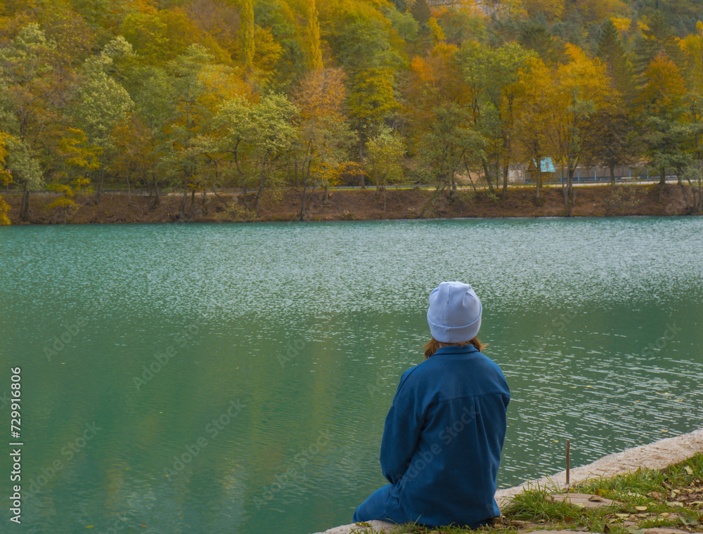 A lonely woman sits on the shore of a mountain forest lake. Autumn landscape, tranquility, tranquility, travel.