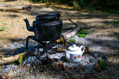 A black kettle and china teapot next to fire in a camp indicating a refreshing vibe photo