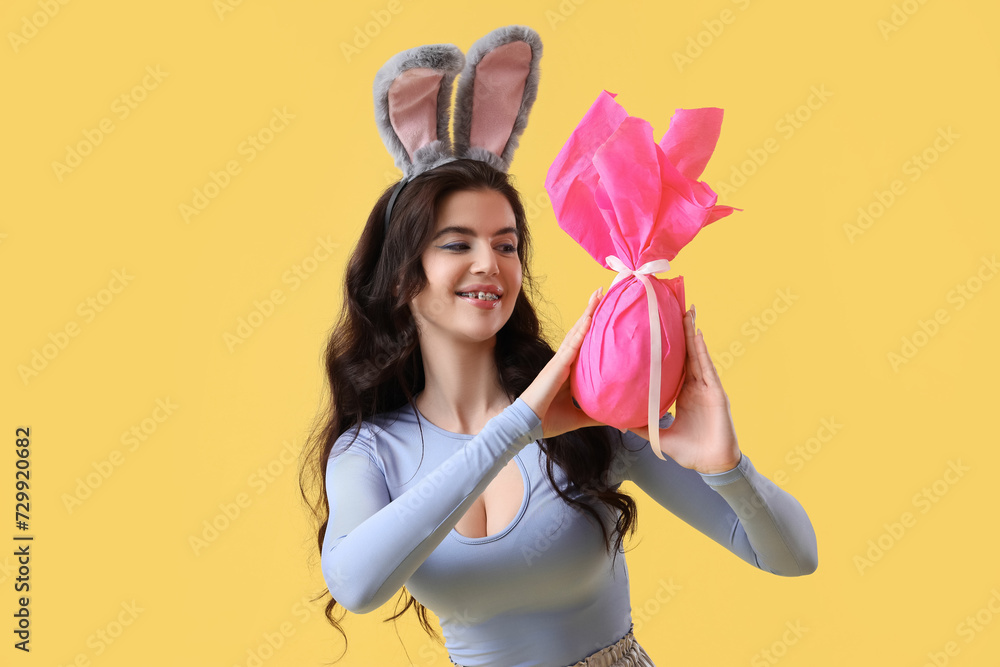 Young woman in bunny ears with Easter gift egg on yellow background Young woman in bunny ears with Easter gift egg on yellow background