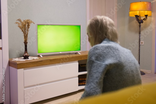 Grandma is watching TV. Green screen. Grandma is sitting on the sofa at home. In front of her is a green screen TV.