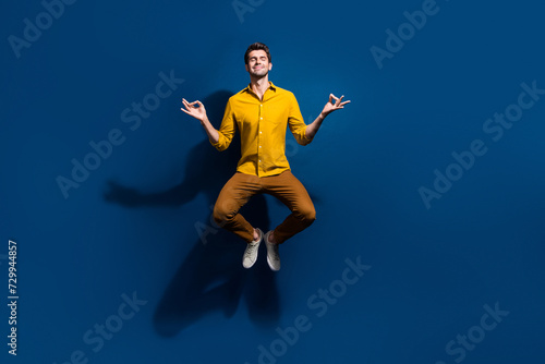 Full size photo of attractive young man jumping meditate closed eyes dressed stylish yellow clothes isolated on dark blue color background