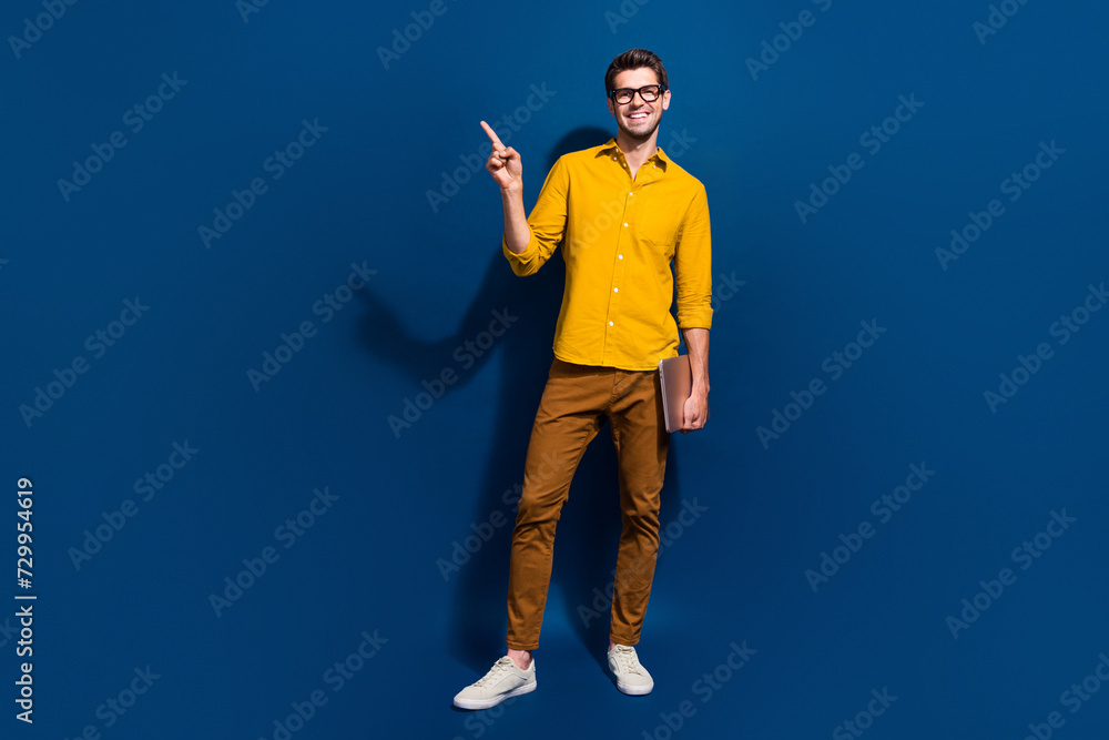 Full length photo of nice young male netbook specs point empty space dressed stylish yellow garment isolated on dark blue color background Full length photo of nice young male netbook specs point empty space dressed stylish yellow garment isolated on dark blue color background