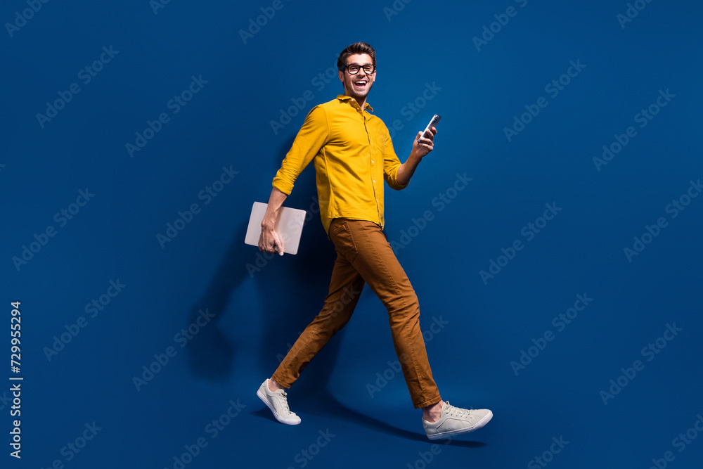 Full body photo of attractive young guy walk netbook device marketer dressed stylish yellow clothes isolated on dark blue color background