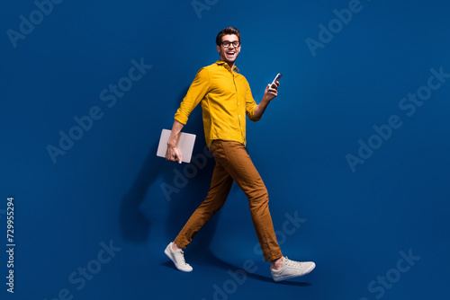 Full body photo of attractive young guy walk netbook device marketer dressed stylish yellow clothes isolated on dark blue color background © deagreez
