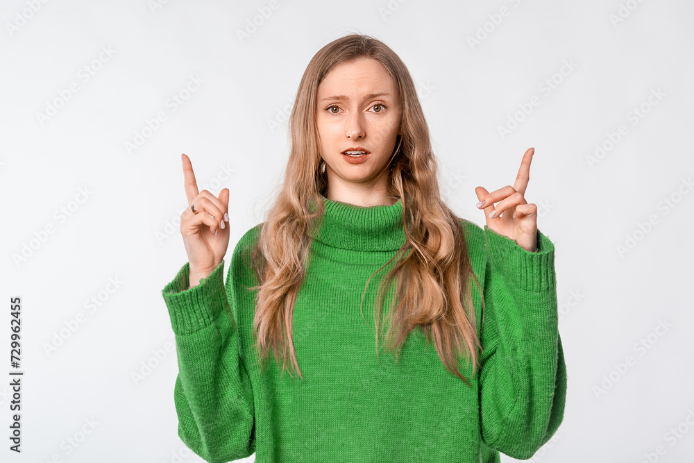 Portrait of surprised woman pointing fingers up, showing advertisement on top, recommending click on link, demonstrating promo banner, wearing green cozy sweater, standing over white background Portrait of surprised woman pointing fingers up, showing advertisement on top, recommending click on link, demonstrating promo banner, wearing green cozy sweater, standing over white background