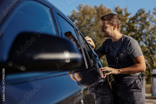 A happy car wash worker is cleaning car window outdoors.