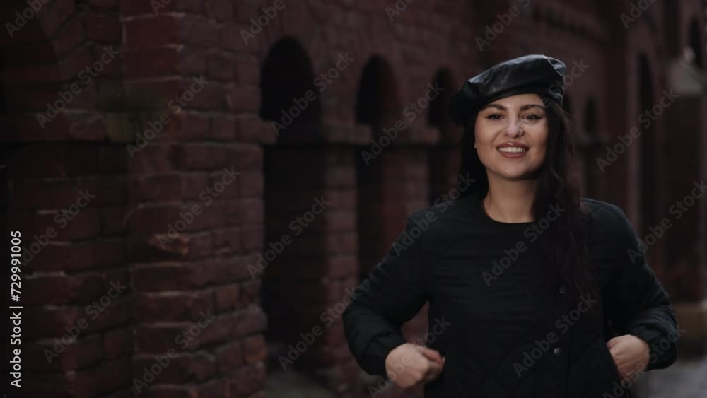 turns around, looking at camera, relaxed lady. Young, cheerful woman in stylish leather cap and quilted jacket stands before aged brick wall, her smile exuding confidence and urban chic.