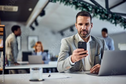 A businessman using a phone while working over the laptop, his colleagues working in the background.