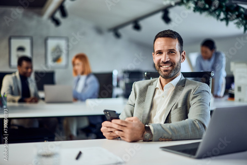 A portrait of a smiling businessman looking at the camera while using a phone and sitting in the co-working space.