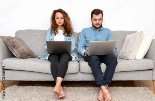serious and focused man and woman sit on sofa next to each other and using their laptops.