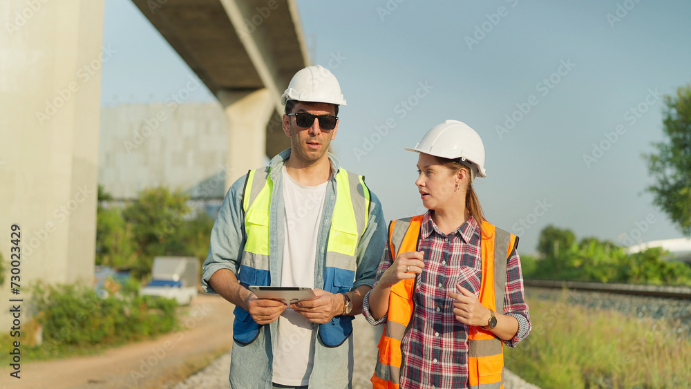 Two engineers in safety uniform and helmet using tablet discussion and inspection construction process railway while walking. Two engineer working on railway together. Teamwork concept Two engineers in safety uniform and helmet using tablet discussion and inspection construction process railway while walking. Two engineer working on railway together. Teamwork concept
