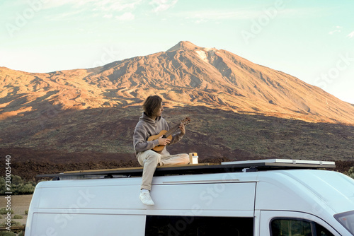 A young man sits on top of a van roof, playing a guitar against the scenic backdrop of Teide Mountain.