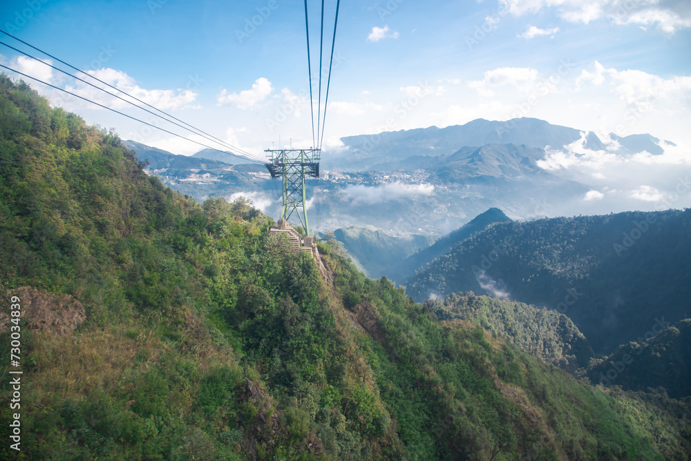 Foggy and sunny scenic view Hoang Lien Son mountain range, Muong Hoa Valley, aerial lift pylon cable car pillar steel framework hauled gondola lift ropeway above ground, cloud blue sky, Sapa