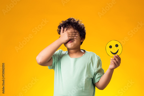 Child boy holds emoticons with happy emotions and closes eyes with palm.