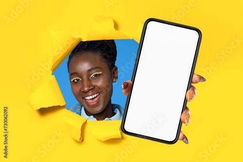 black woman showing phone screen through hole in yellow background © Prostock-studio