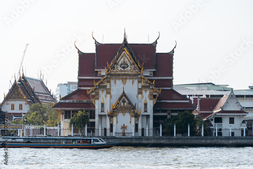 A traditional Thai temple by the riverside with a boat passing by in Bangkok.
