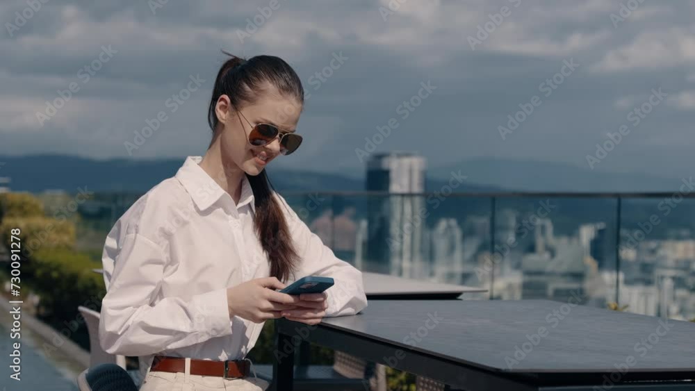 Business woman in sunglasses with a smile looking into the screen of the cell phone in her hands outside in the sun on the background of the city and skyscrapers in the afternoon, lifestyle urban Business woman in sunglasses with a smile looking into the screen of the cell phone in her hands outside in the sun on the background of the city and skyscrapers in the afternoon, lifestyle urban