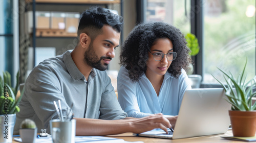 Two professionals are closely working together on a project, looking at a laptop screen in a modern office setting.