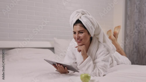 Technology leisure, Fruit enjoyment, Comfortable position. Lying prone in bedroom, woman enjoys free time by using tablet and savoring apple.