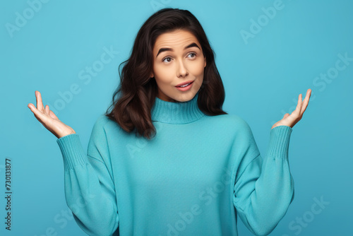 Young woman with arms out, shrugging her shoulders, saying: who cares, so what, I don't know. Isolated studio shot on blue background. Whatever gesture. photo