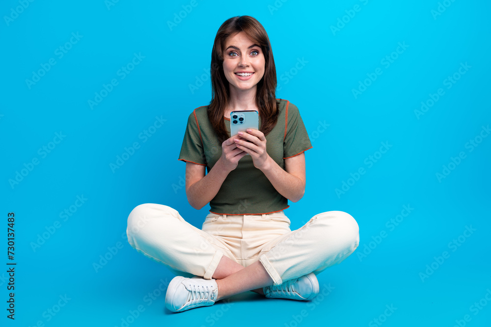 Full size photo of cheerful woman wear green t-shirt sit on floor hold smartphone read notification isolated on blue color background Full size photo of cheerful woman wear green t-shirt sit on floor hold smartphone read notification isolated on blue color background