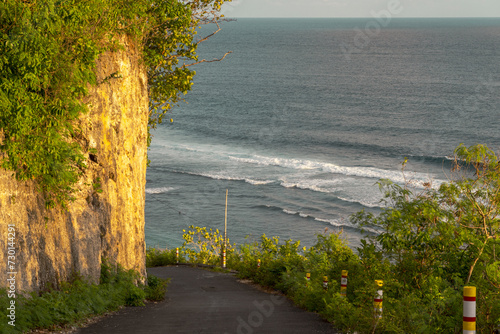The road bends dramatically, revealing an expansive ocean vista. The golden hour sunlight bathes the scene in a warm glow, highlighting the natural beauty of the coastal landscape.