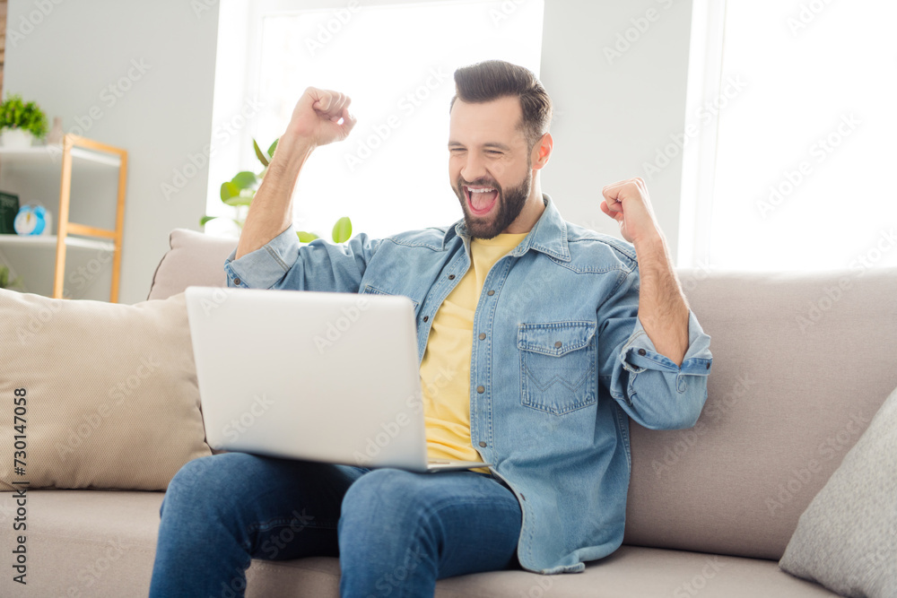 Portrait of astonished cheerful man sit on sofa look laptop fists up open mouth shout yes free time home indoors Portrait of astonished cheerful man sit on sofa look laptop fists up open mouth shout yes free time home indoors