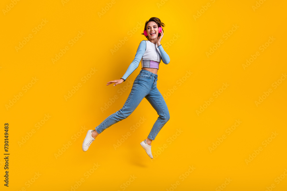 Full size portrait of overjoyed girl arm touch headphones jump empty space ad isolated on yellow color background
