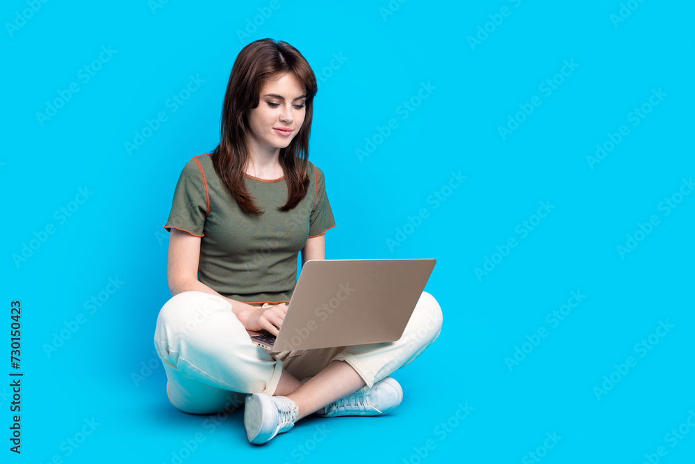 Full size photo of focused intelligent woman wear green t-shirt sit on floor typing message on laptop isolated on blue color background Full size photo of focused intelligent woman wear green t-shirt sit on floor typing message on laptop isolated on blue color background