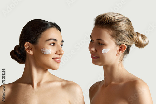 Blonde and brunette beautiful girls with clear, well-kept skin applying face moisturizing cream against white studio background. Concept of natural beauty, cosmetology and cosmetics, skincare