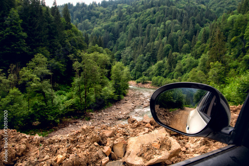 View of a car and a car mirror towards a river in a mountainous area. Landslides and consequences of mountain river flooding.
