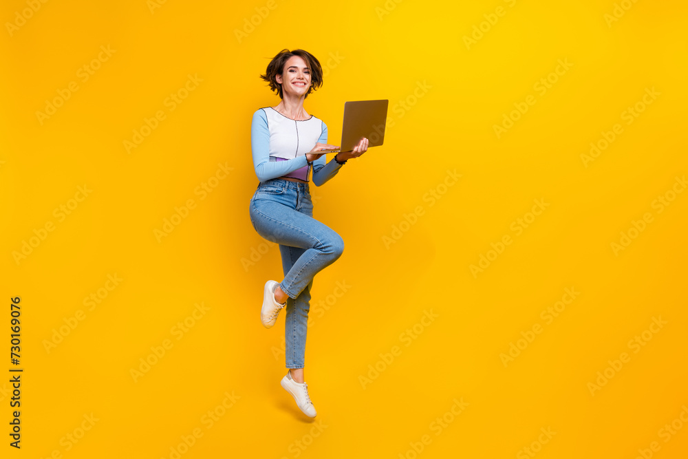 Full size portrait of gorgeous girl jump hold wireless laptop empty space isolated on yellow color background Full size portrait of gorgeous girl jump hold wireless laptop empty space isolated on yellow color background