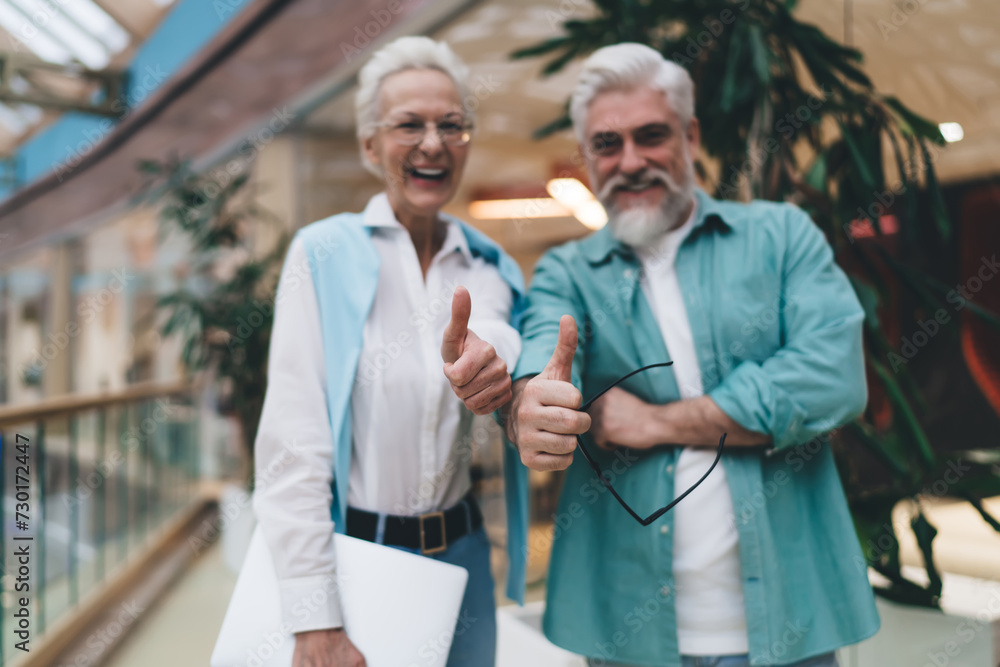 Obraz premium Cheerful senior Caucasian businesswoman and businessman giving thumbs up in a bright office atrium, celebrating success or agreement, showcasing teamwork and positive corporate culture