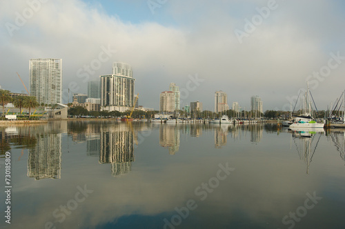 Wide shot Looking north from Demens Landing Park over smooth water with reflections towards city scape of St. Petersburg  Florida . Sailbots in marina with Cloudy blue and white sky with light fog.