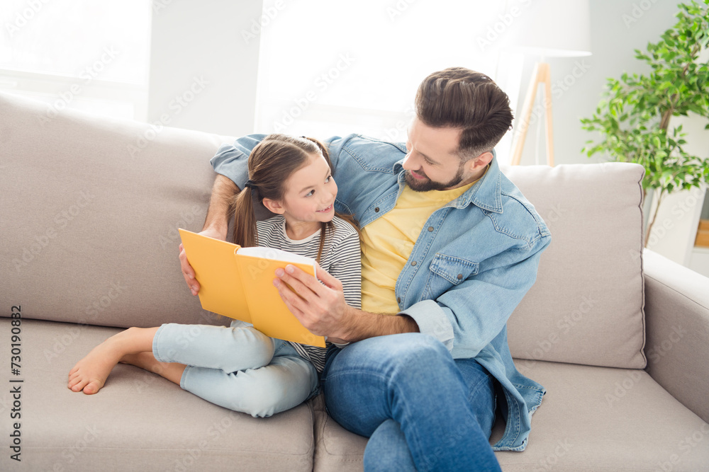 Portrait of cheerful little girl positive guy sit on sofa hold book look each other have good mood free time indoors Portrait of cheerful little girl positive guy sit on sofa hold book look each other have good mood free time indoors