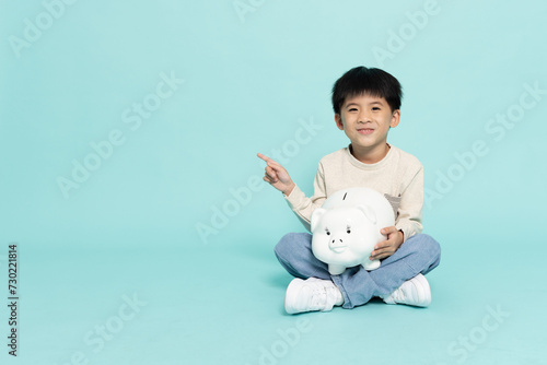 Asian little boy sitting on floor and pointing to copy space with holding white piggy bank isolated on green background, Saving money and financial economy concept