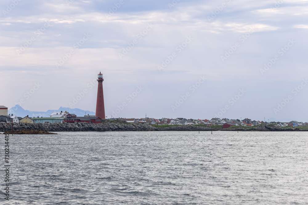 A towering red lighthouse, a beacon of Andenes, Norway, rises above a calm sea with a backdrop of a cozy, colorful town.