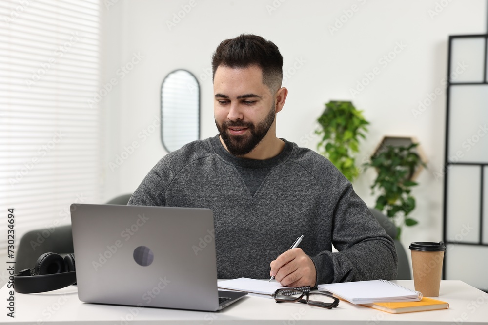 E-learning. Young man taking notes during online lesson at white table indoors E-learning. Young man taking notes during online lesson at white table indoors