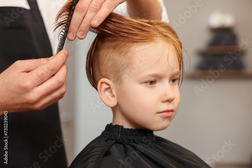 Professional hairdresser combing boy's hair in beauty salon, closeup