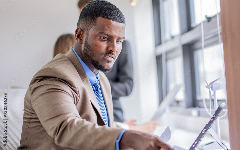 Fototapeta premium Selective focus African smart handsome businessman, wearing formal suit and necktie, working, reading project plan of green eco friendly project seriously, sitting in workplace in indoor office.