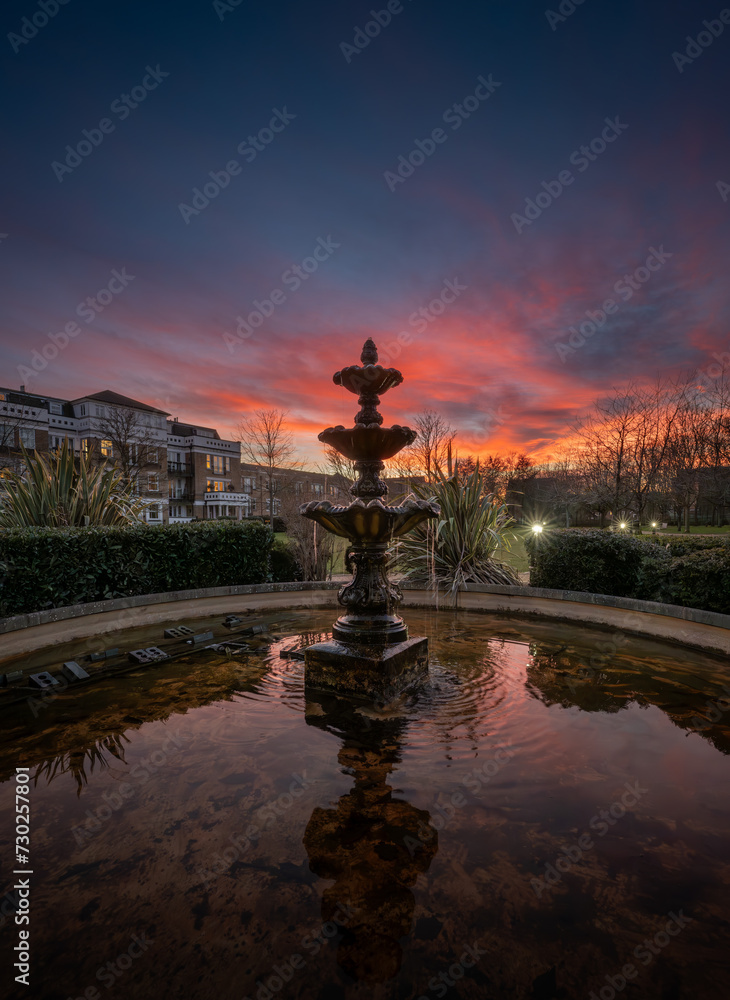 Fountain and pond in a park at sunset with a pink sky. Evening view in portrait orientation with buildings and trees behind.