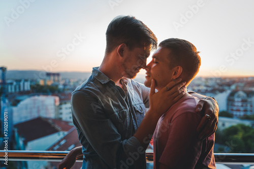 Young happy cute gay couple hugging and kissing on balcony overlooking city and sunset