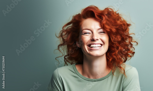 Joyful mature woman with curly red hair and radiant smile  wearing a green top against a light grey background