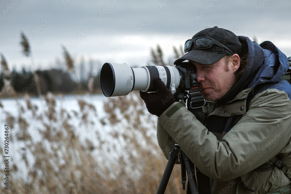 Wildlife photographer is setting up camera on tripod outdoors. Man photographing landscape or animals at lake in winter. Bird watching