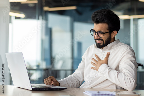 Overworked indian businessman feeling unwell at desk with laptop in modern office photo