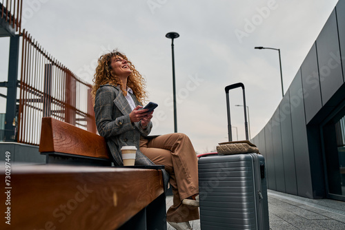 Sitting on a bench, a Caucasian woman uses her phone, holding a takeaway coffee with a suitcase beside her, patiently waiting for the bus. photo