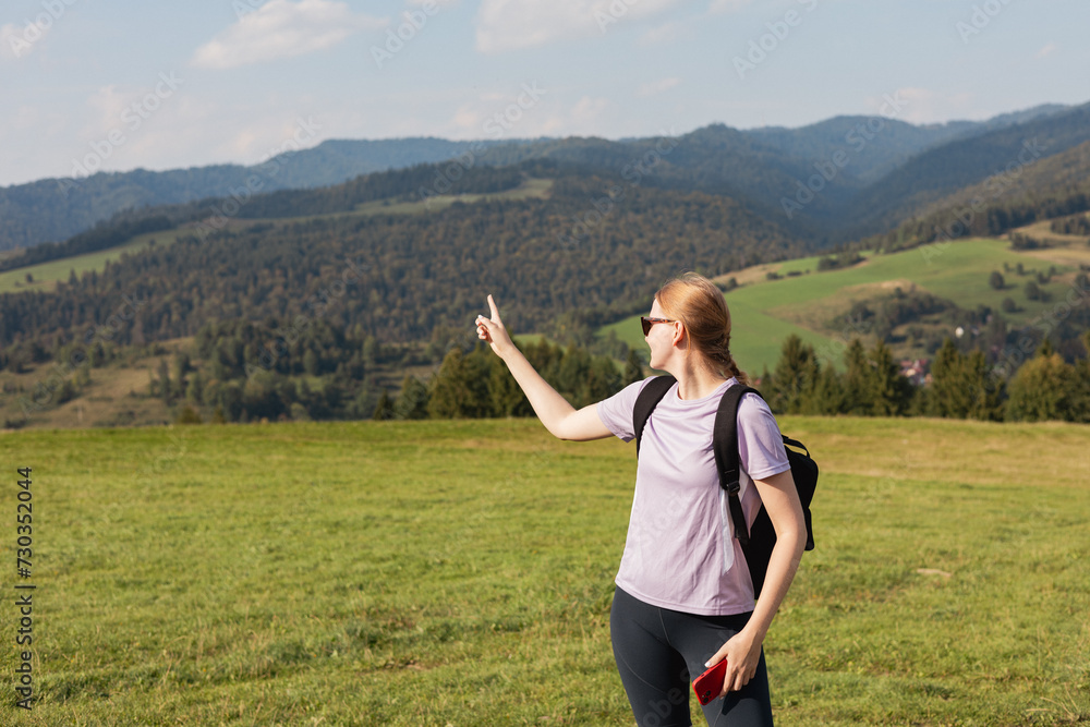 Blonde 30s girl in sunglasses pointing finger on mountains background. Young female with backpack hiking in nature. Beautiful woman walking in sport clothes. Freedom and active lifestyle concept