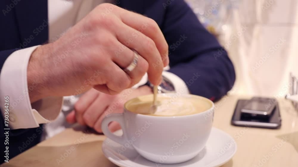 Male hand with ring stirs coffee cappuccino in cafe, slow motion, smartphone out of focus Male hand with ring stirs coffee cappuccino in cafe, slow motion, smartphone out of focus